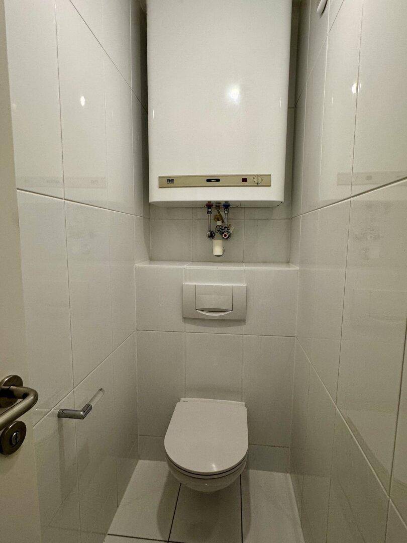 Kleines, gefliestes WC mit wandhängender Toilette und einem Warmwasserboiler an der Wand.