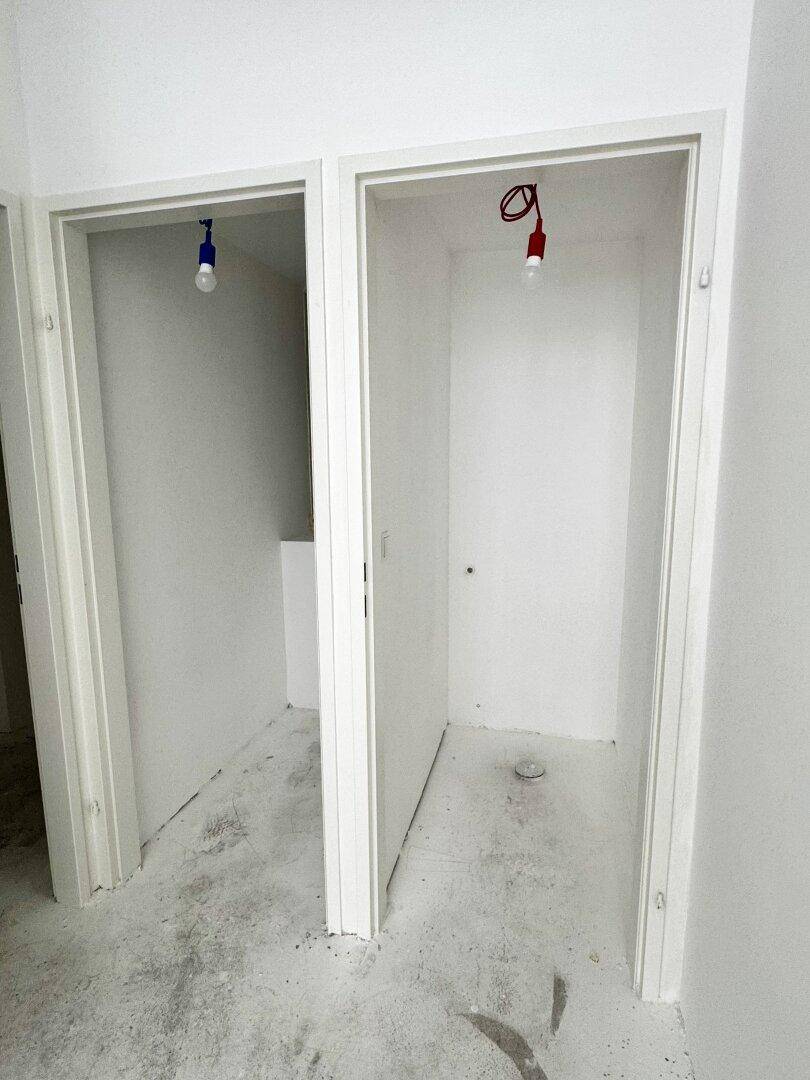 Zwei offene Türrahmen führen zu kleinen, leeren Räumen mit Betonboden und weißen Wänden, möglicherweise Abstellräume.