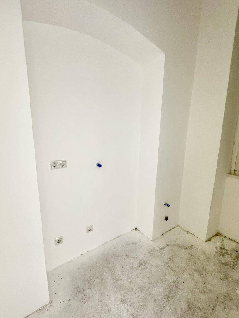 Ecke eines leeren Raumes mit weißen Wänden, sichtbaren Steckdosen und Wasseranschlüssen für zukünftige Installationen.