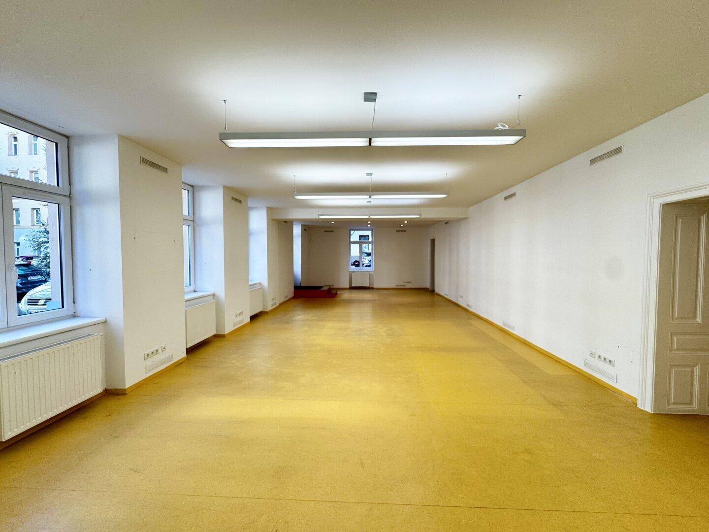 Langer, heller Büroraum mit gelbem Boden, weißen Wänden und mehreren Fenstern zur Straße.
