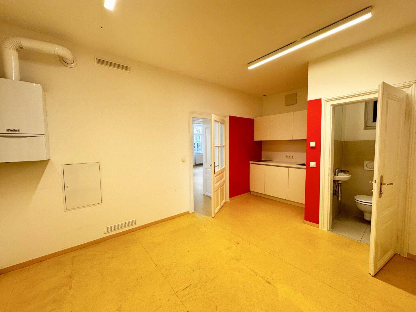 Raum mit kleiner Küchenzeile, roter Akzentwand und Zugang zu einer separaten Toilette.