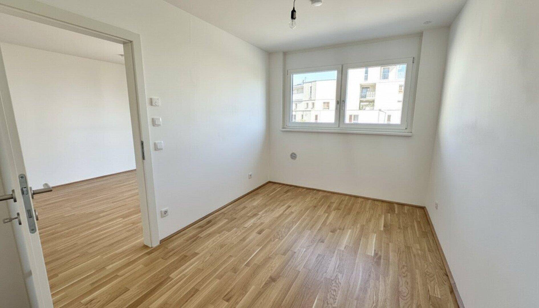 Leeres Schlafzimmer mit Parkettboden und einem Fenster, das viel Tageslicht hereinlässt.
