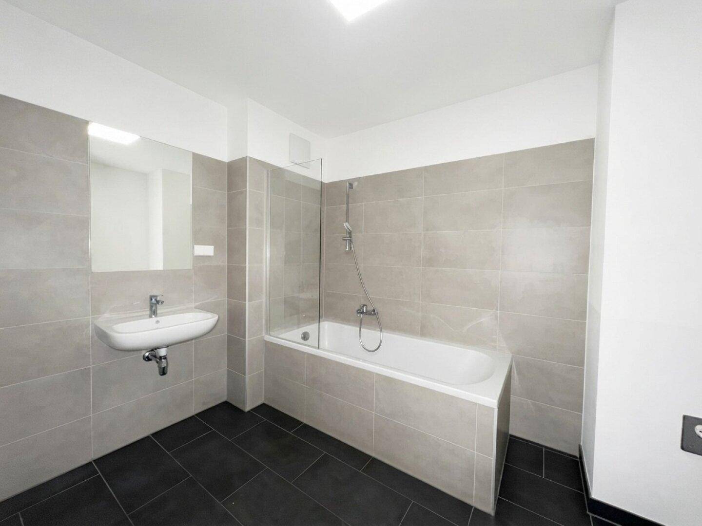 Modernes Badezimmer mit Badewanne, Waschbecken und grauen Wand- und dunklen Bodenfliesen.
