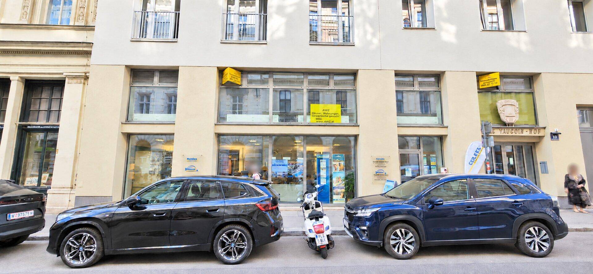 Ansicht der Gebäudefassade mit großen Fenstern, Geschäften im Erdgeschoss und parkenden Fahrzeugen.
