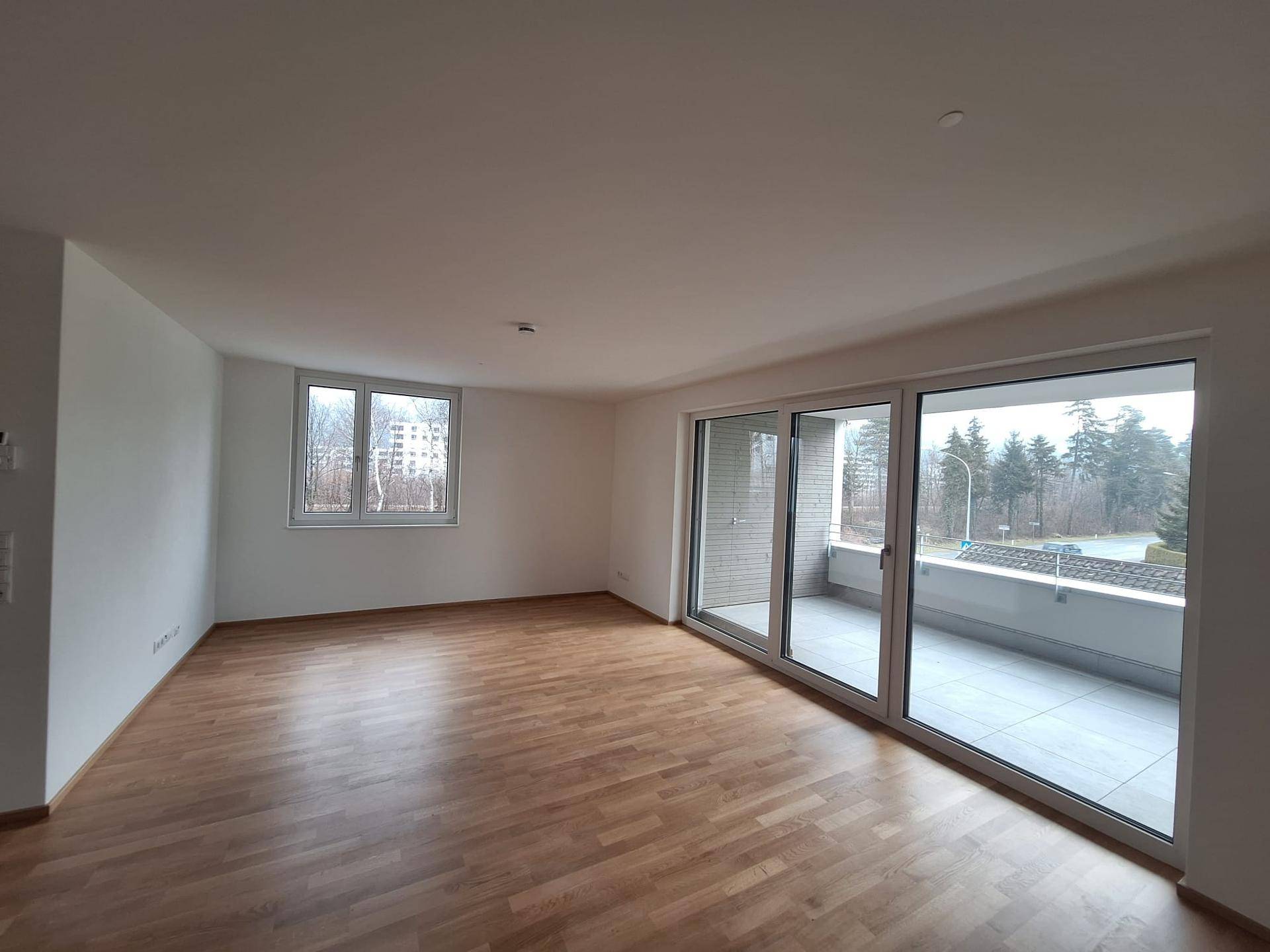 Geräumiger Wohnbereich mit großen Fenstern, Holzboden und direktem Zugang zum Balkon.