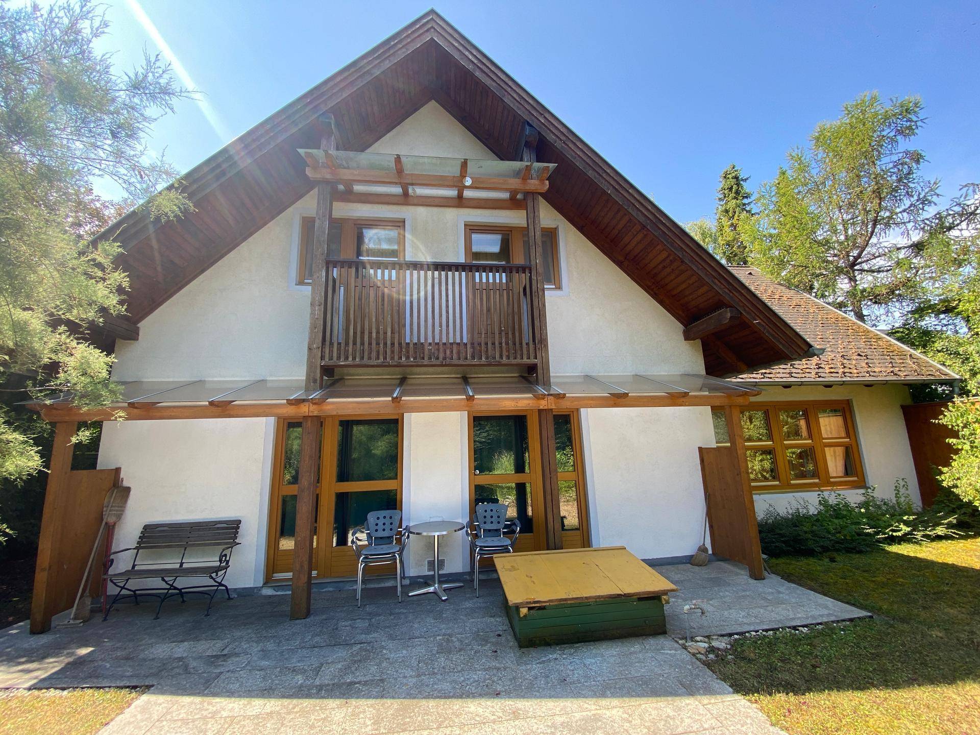 Hausfassade mit Giebeldach, Holzbalkon und kleiner Terrasse mit Sitzgelegenheiten im Freien.