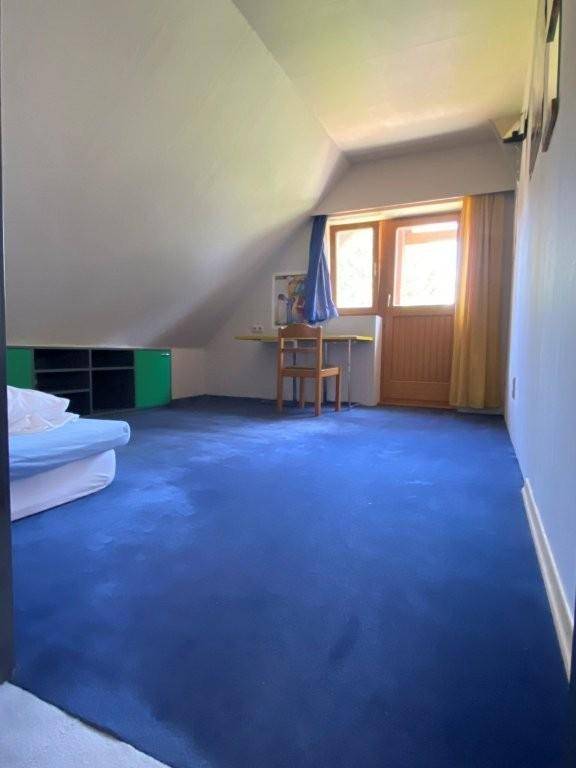 Schlafzimmer im Dachgeschoss mit blauem Teppichboden, Schreibtisch und Fenster mit Blick ins Grüne.