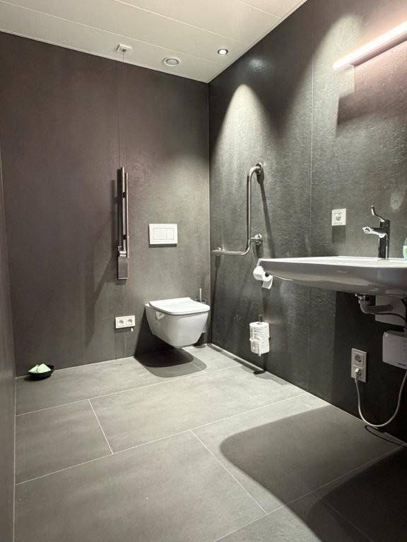 Modernes barrierefreies Badezimmer mit dunklen Fliesen, Hänge-WC, Waschbecken und Haltegriffen.