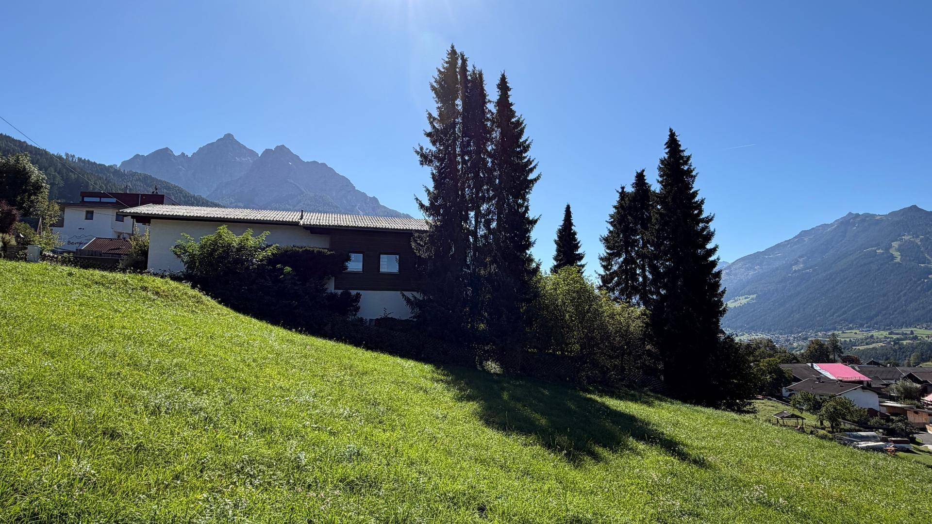 Haus auf einem grünen Hügel mit Blick auf die umliegende Berglandschaft an einem sonnigen Tag.