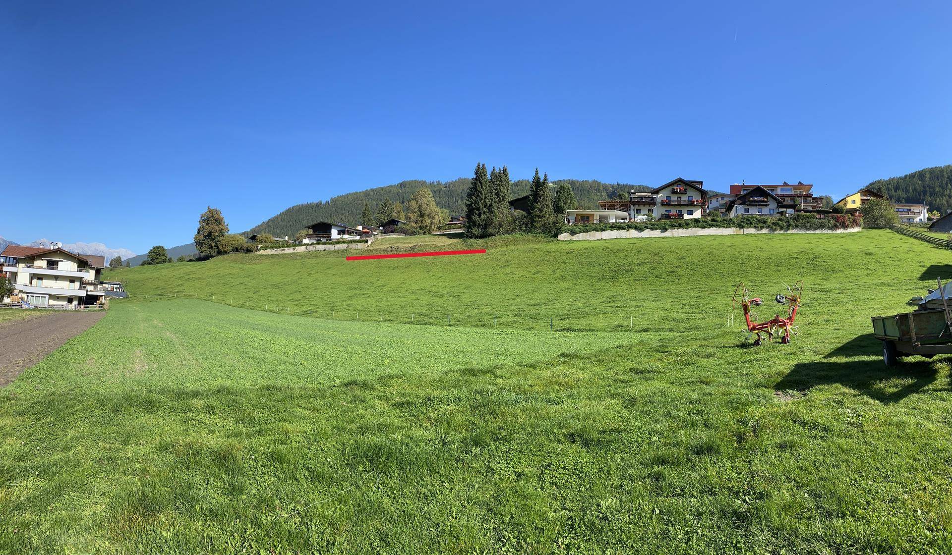 Weitläufige grüne Wiese mit Häusern am Horizont und landwirtschaftlichen Geräten im Vordergrund.