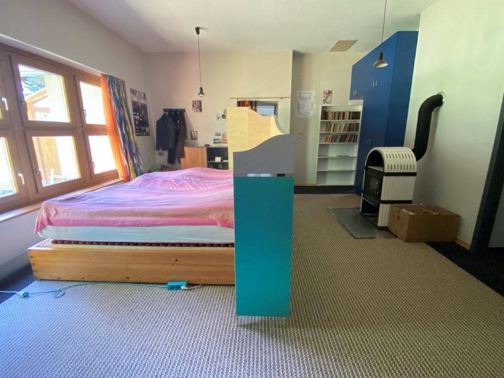 Schlafzimmer mit Holzbett, Teppichboden und einem modernen Holzofen, hell durch große Fenster.