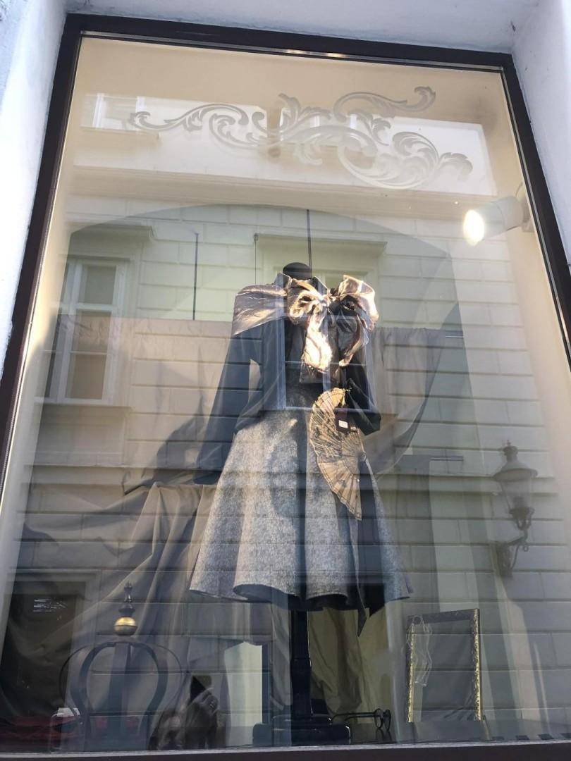 Schaufenster mit einer weiteren eleganten Schaufensterpuppe, die ein graues Kleid und eine große Schleife trägt, reflektiert die Umgebung.