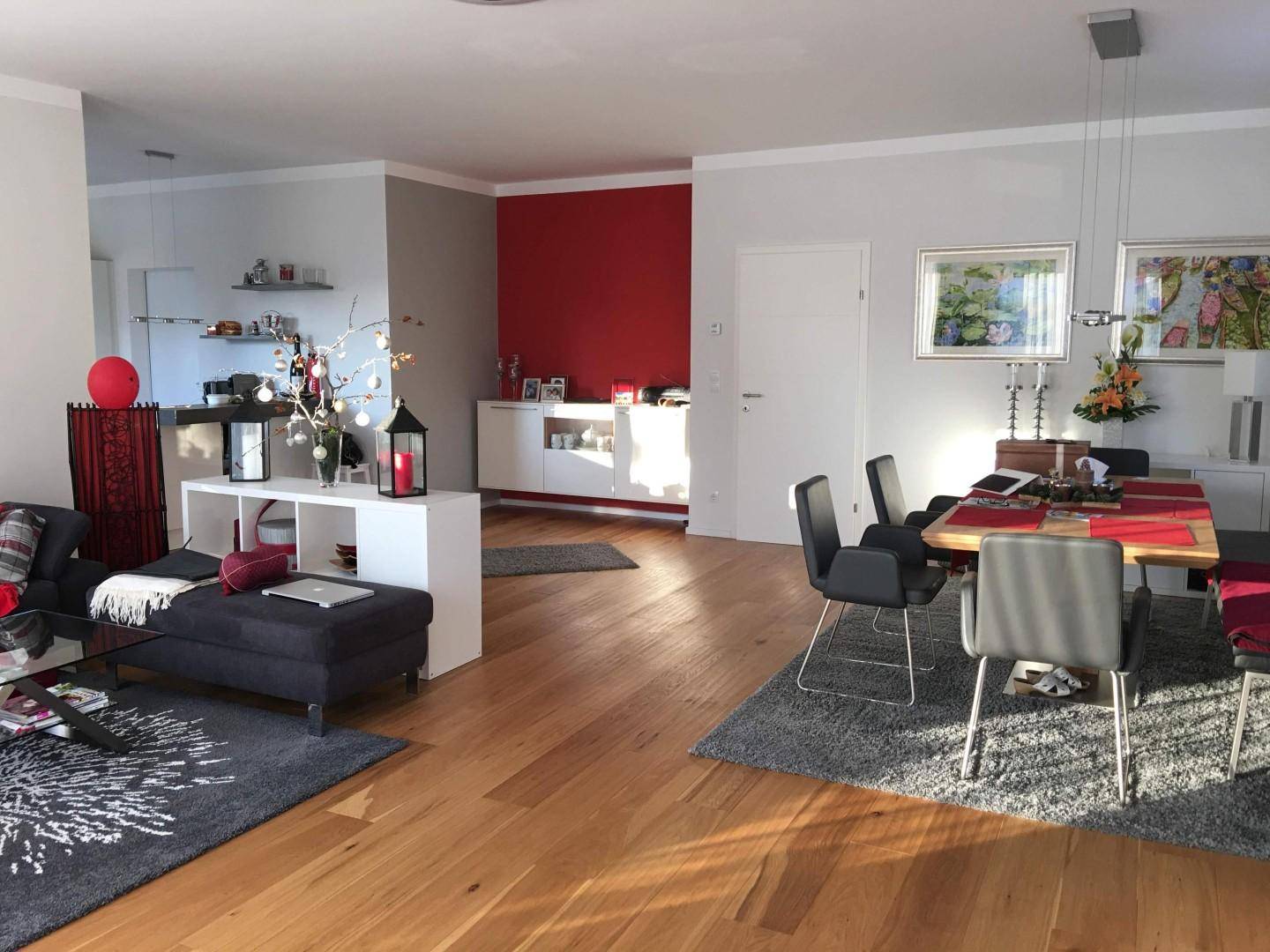 Weitläufiger Wohn- und Essbereich mit Holzboden, roter Akzentwand und viel Tageslicht.