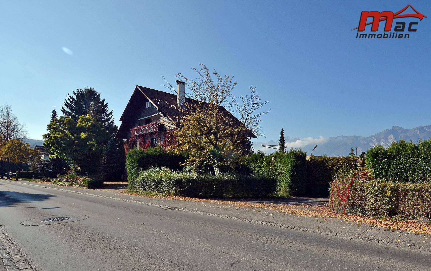 Seitenansicht des Hauses mit gepflegter Hecke und Blick auf die umliegende Berglandschaft im Sonnenschein.