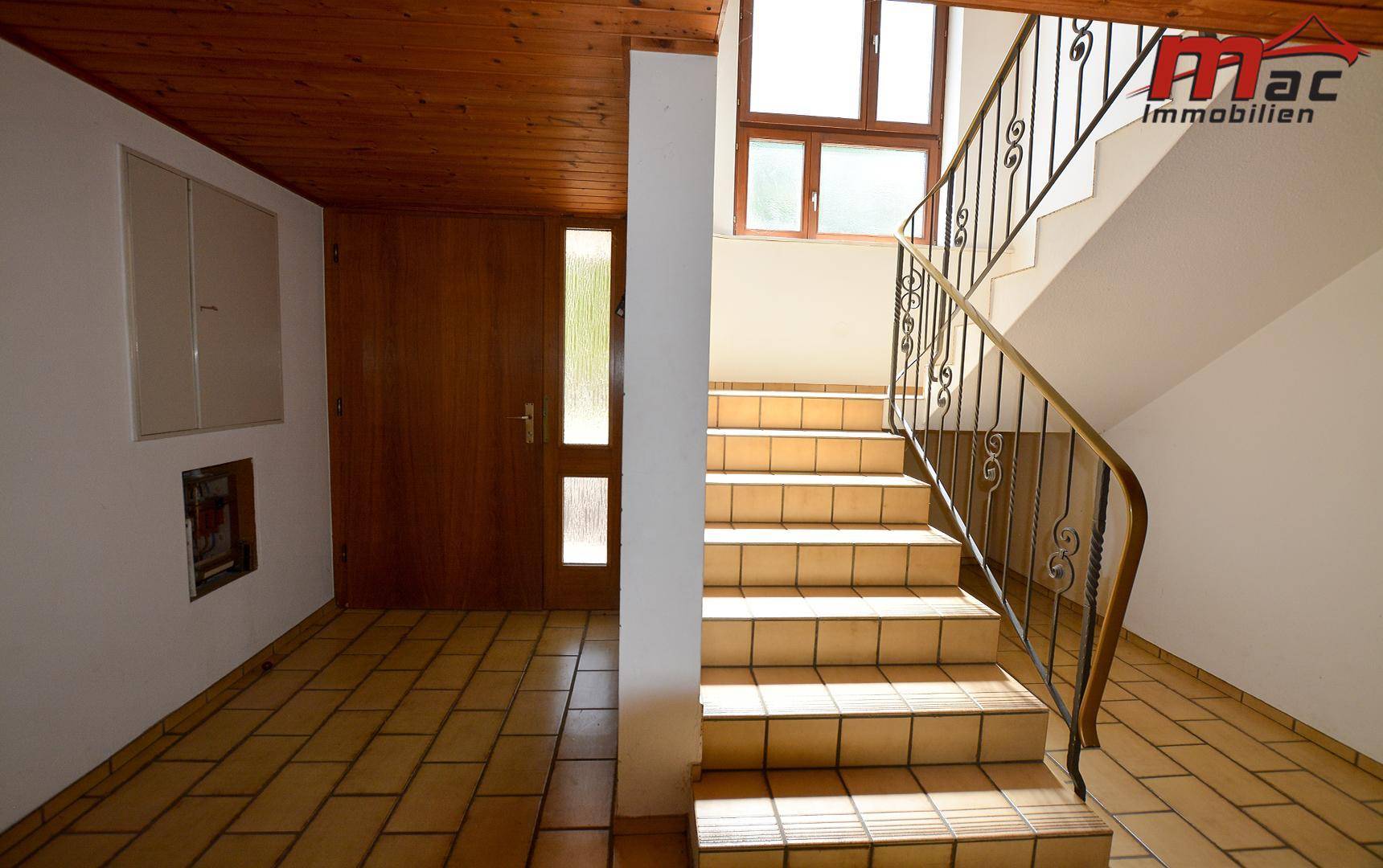 Heller Eingangsbereich mit gefliestem Boden, Holzdecke und einer geschwungenen Treppe mit schmiedeeisernem Geländer.