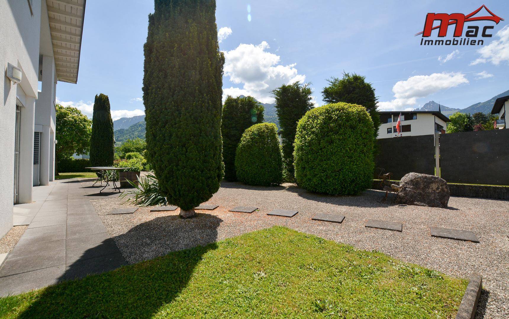 Weitläufiger Garten mit Kiesflächen, Rasen und gepflegten Hecken vor Bergkulisse.