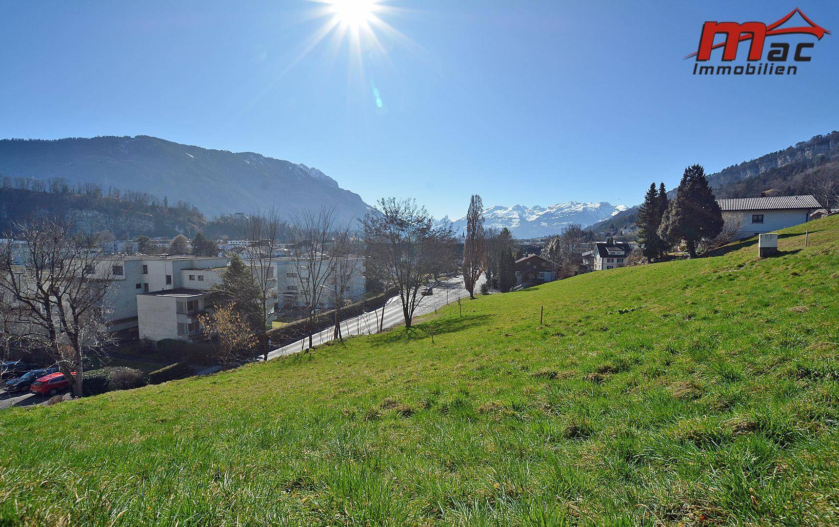 Weitläufige grüne Wiese mit Blick auf eine Siedlung und die majestätischen Alpen im Hintergrund an einem klaren Tag.