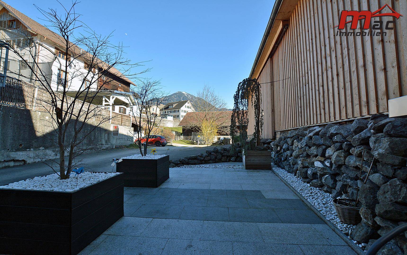 Gepflasterter Außenbereich mit Steinmauer, Pflanzenkübeln und Zugang zum Gebäude.