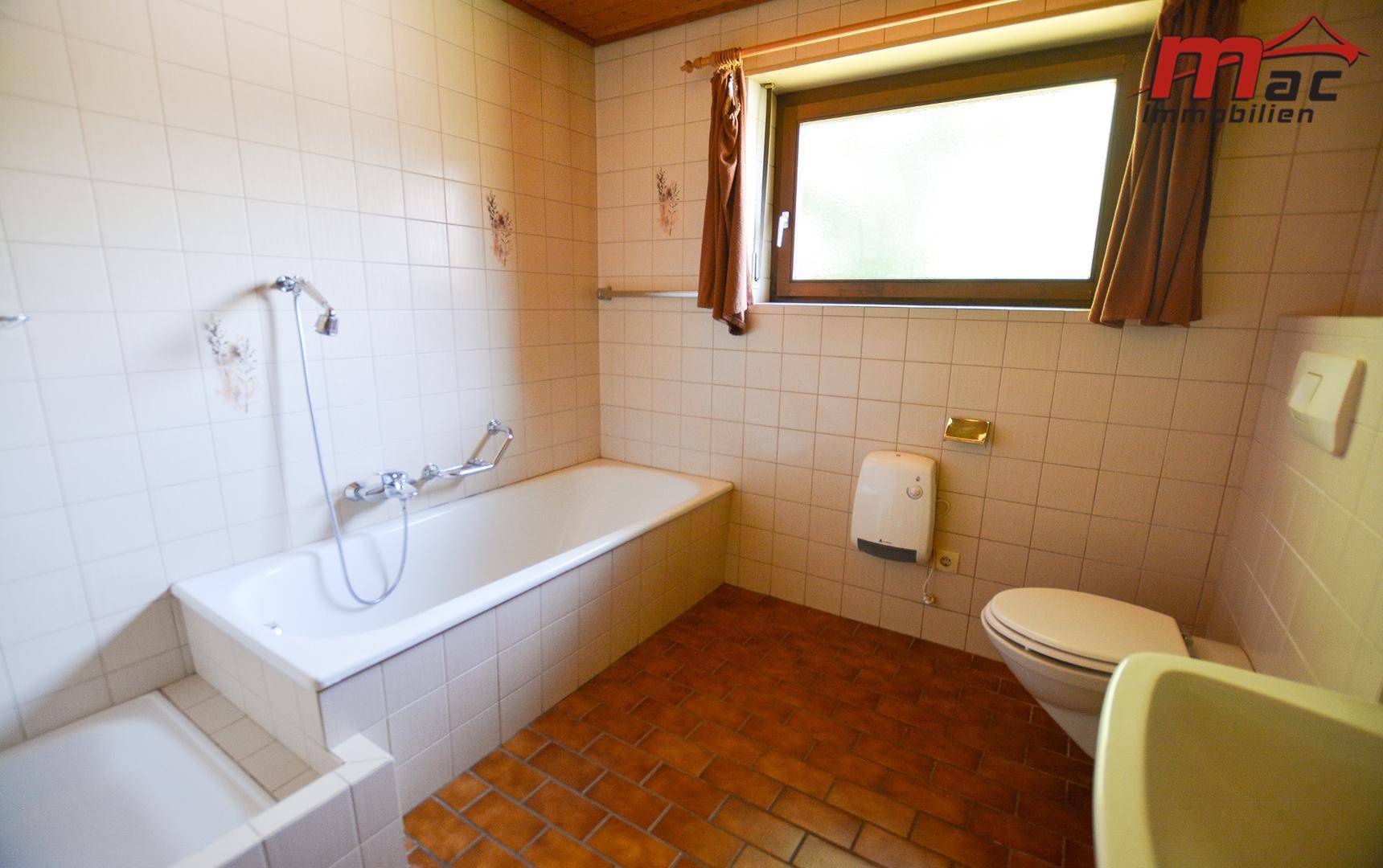 Helles Badezimmer mit Badewanne, Toilette, Waschbecken und terracottafarbenen Bodenfliesen.