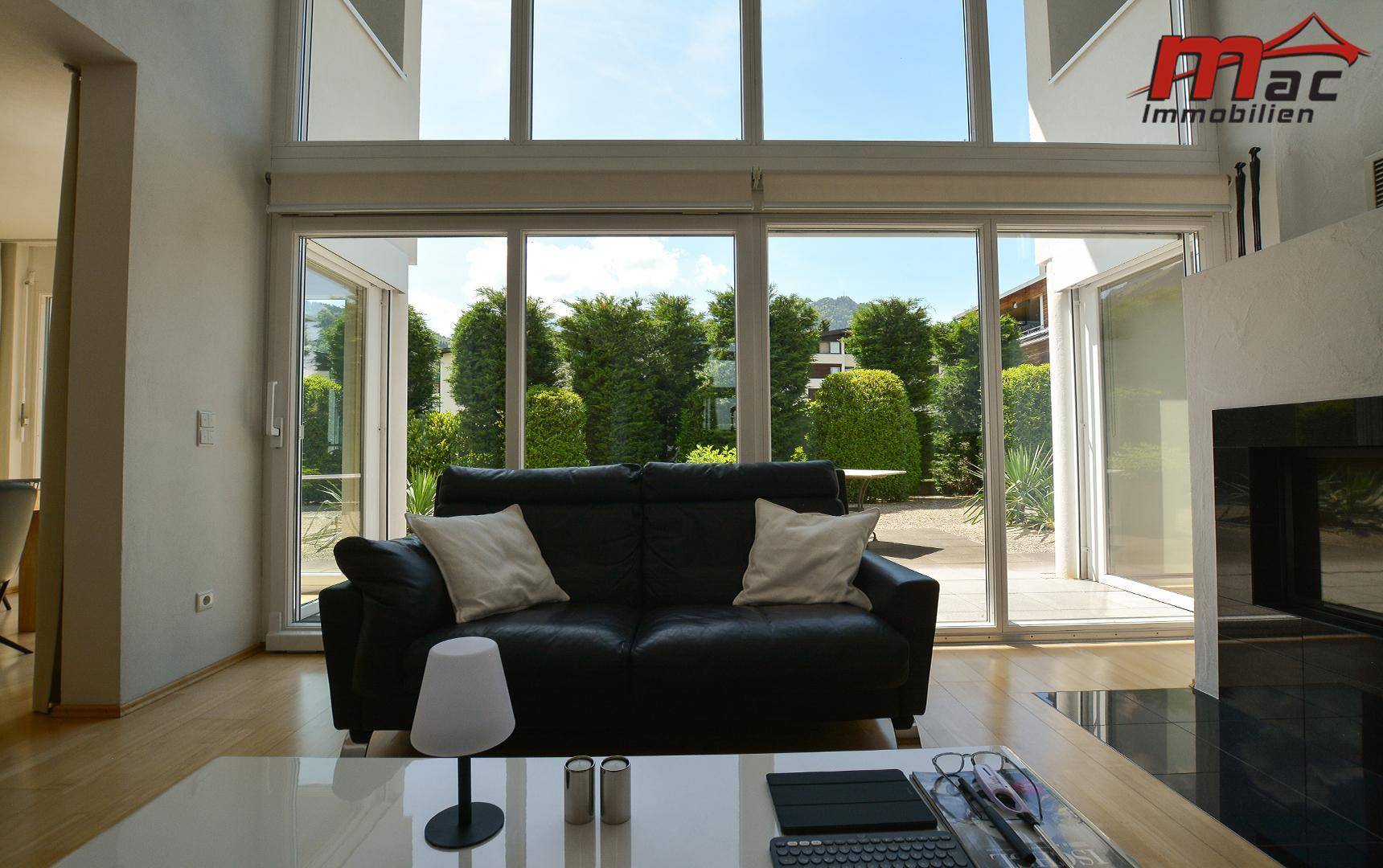 Helles Wohnzimmer mit schwarzem Sofa, Kamin und großen Fensterfronten zum grünen Garten.