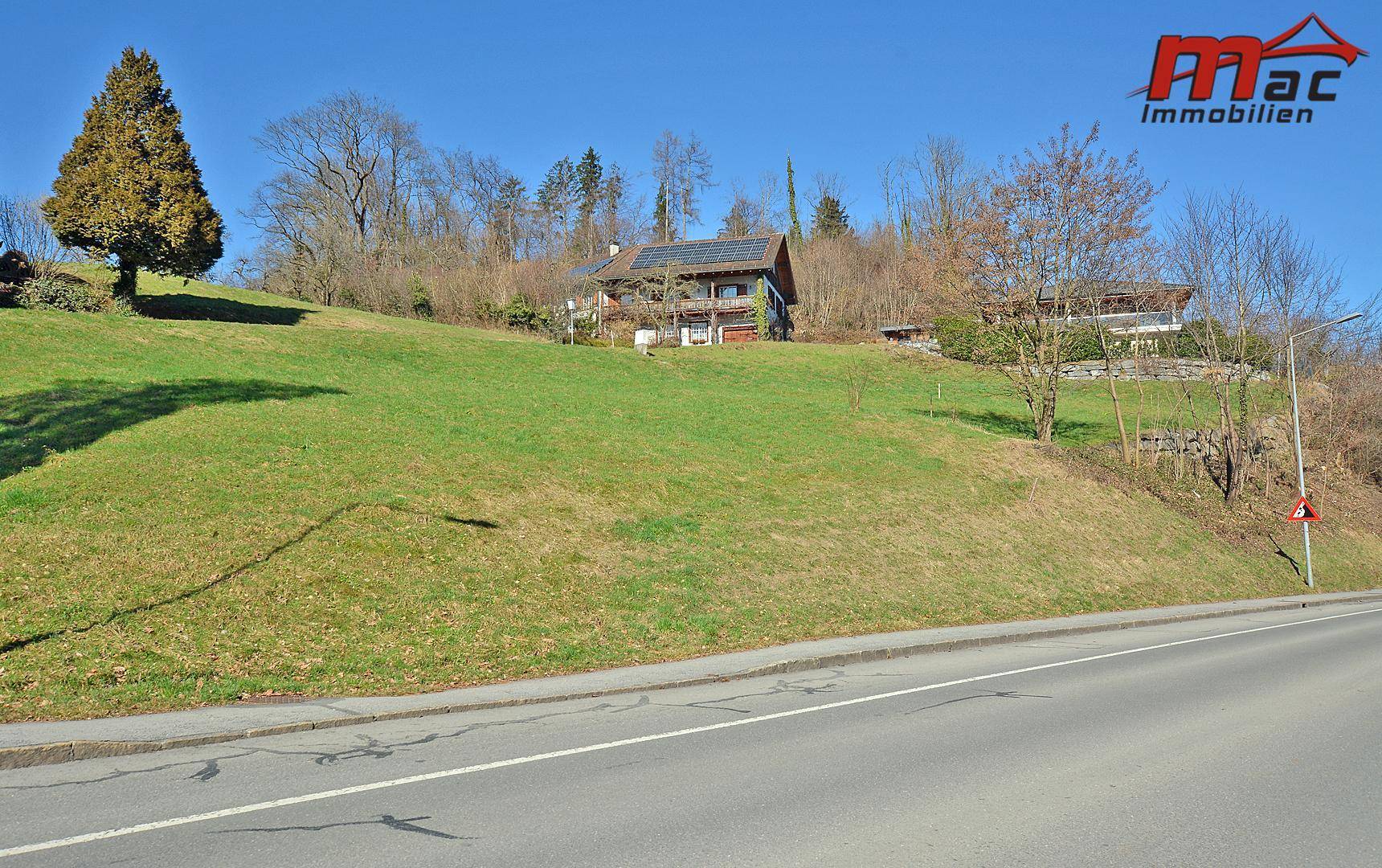 Grüne Hanglage mit einer Straße im Vordergrund und einem Haus mit Solaranlage auf dem Dach in der Ferne.