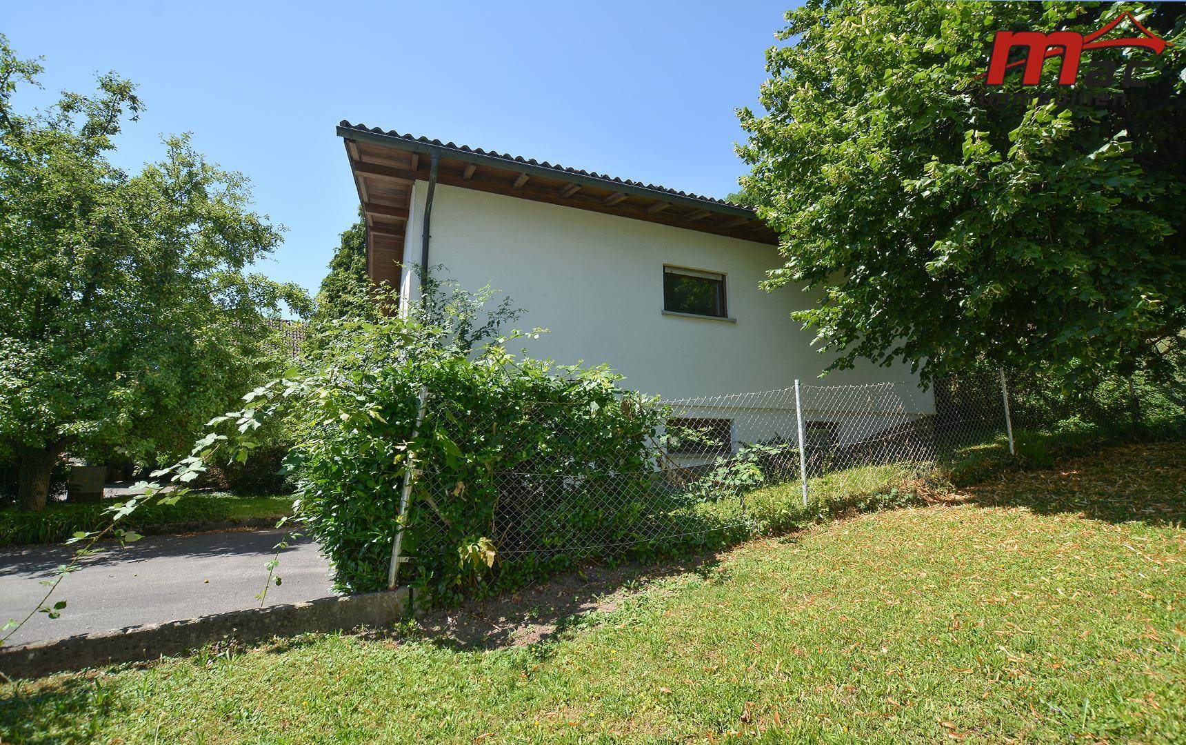 Seitenansicht eines Hauses mit weißer Fassade, umgeben von Bäumen und Grünflächen.