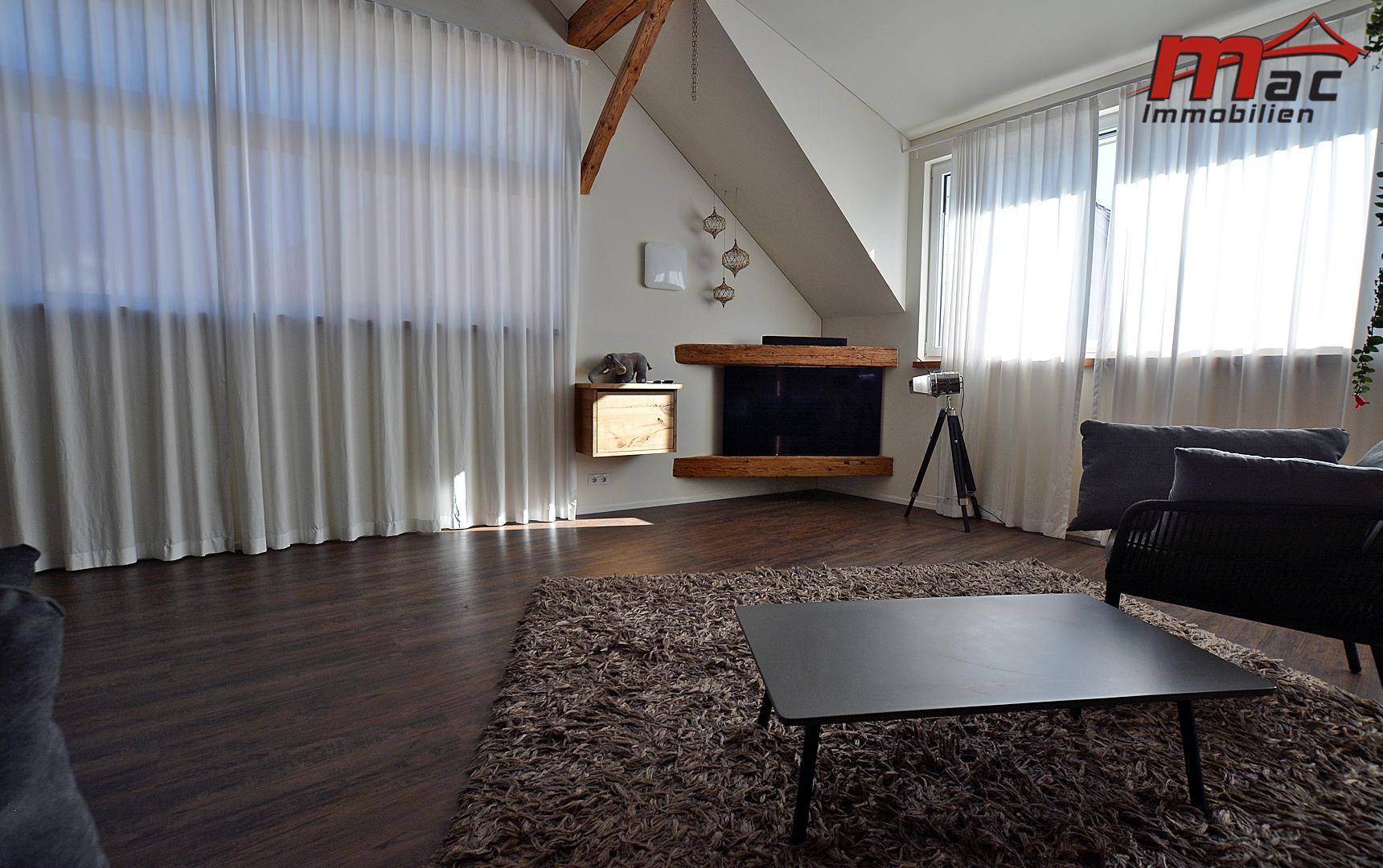 Helles Wohnzimmer mit großen Fenstern, dunklem Holzboden und gemütlichem Teppich.