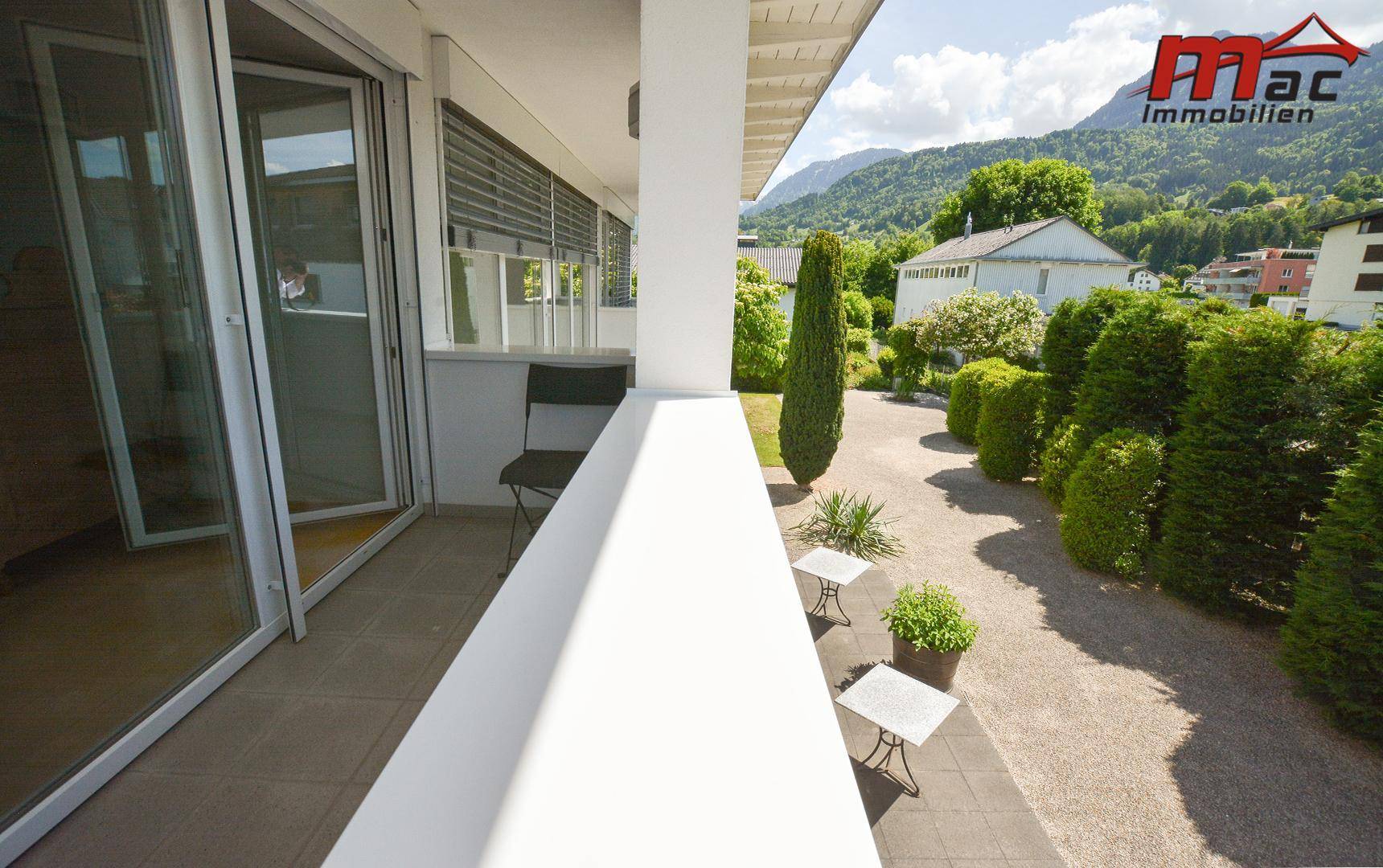 Langer Balkon mit Fliesenboden, Glasfronten und Blick auf den Garten und die Berge.