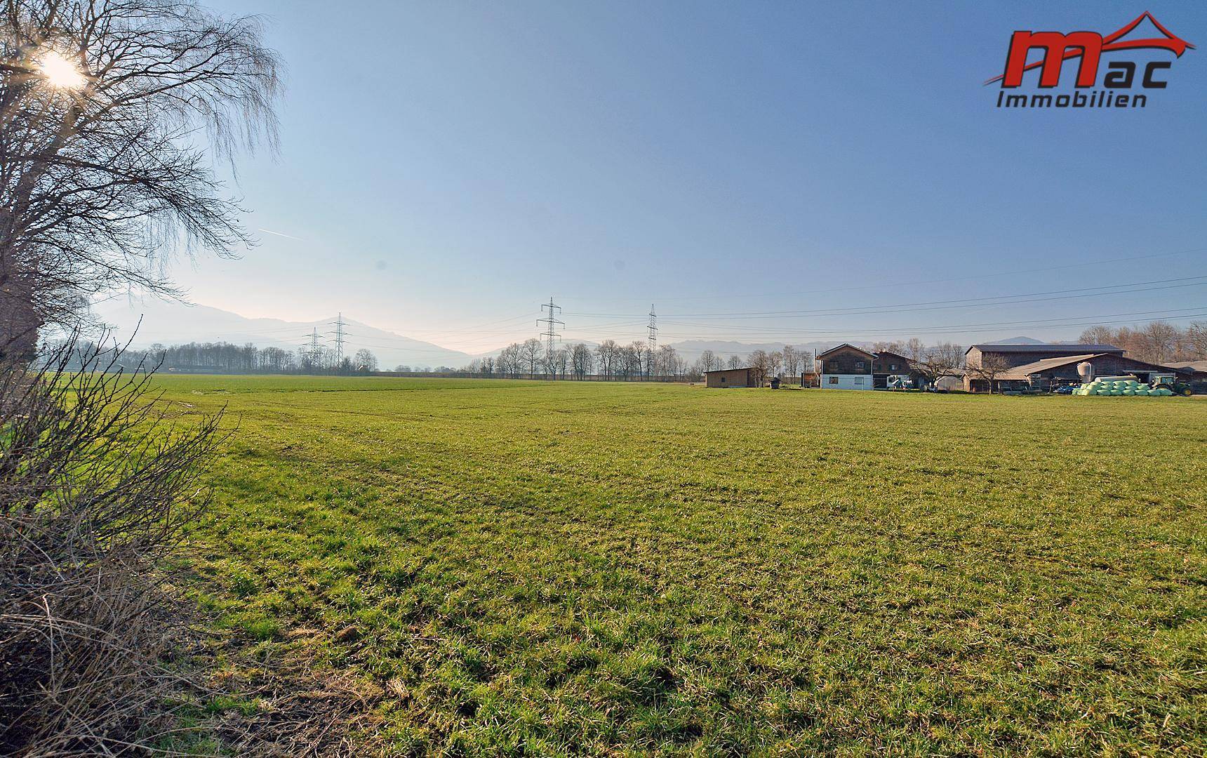Weitläufige grüne Felder mit Bergen im Hintergrund und Stromleitungen am Horizont.