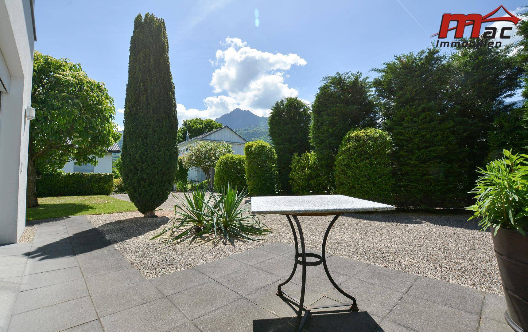 Sonnige Terrasse mit kleinem Tisch und Blick auf den gepflegten Garten und die Berge.