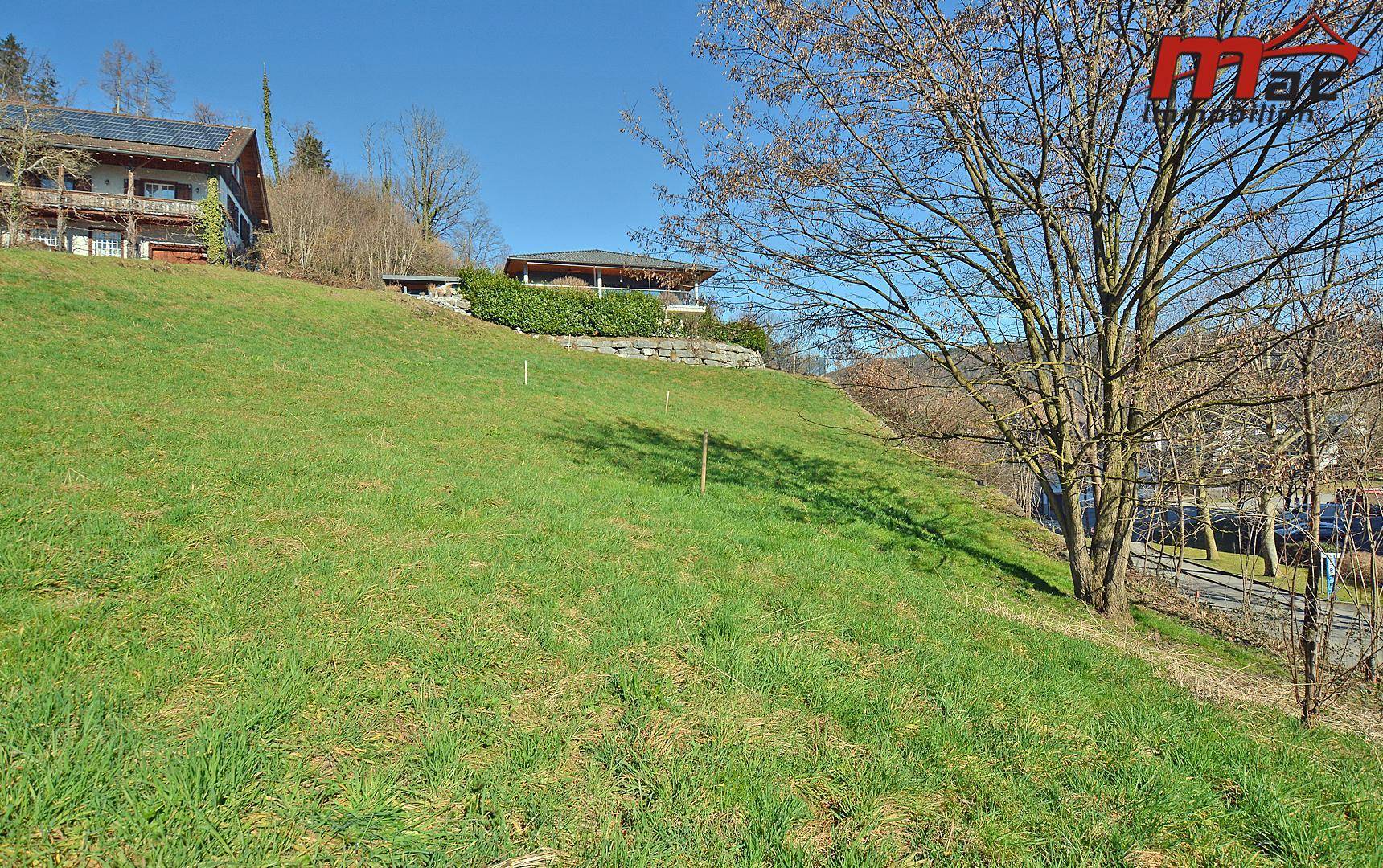 Sonnige Hanglage mit einer grünen Wiese, einem Haus mit überdachter Terrasse und einer Straße im Vordergrund.
