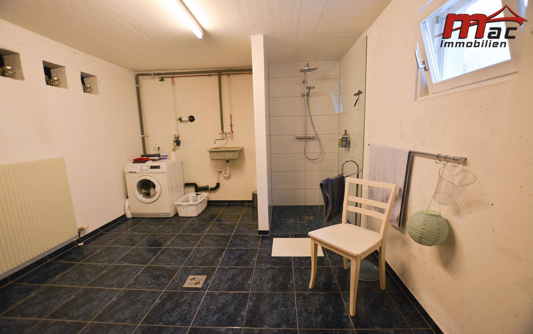 Praktischer Waschraum mit Dusche, Waschmaschine und dunklen Bodenfliesen im Kellerbereich.