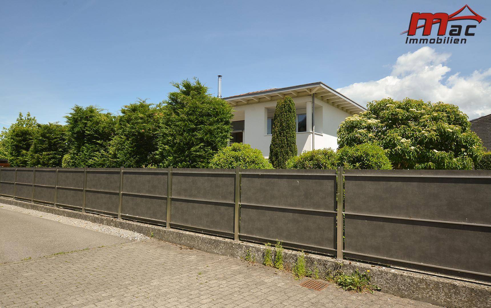 Ansicht des Hauses von der Straße, umgeben von hohen Bäumen und einem modernen Zaun.