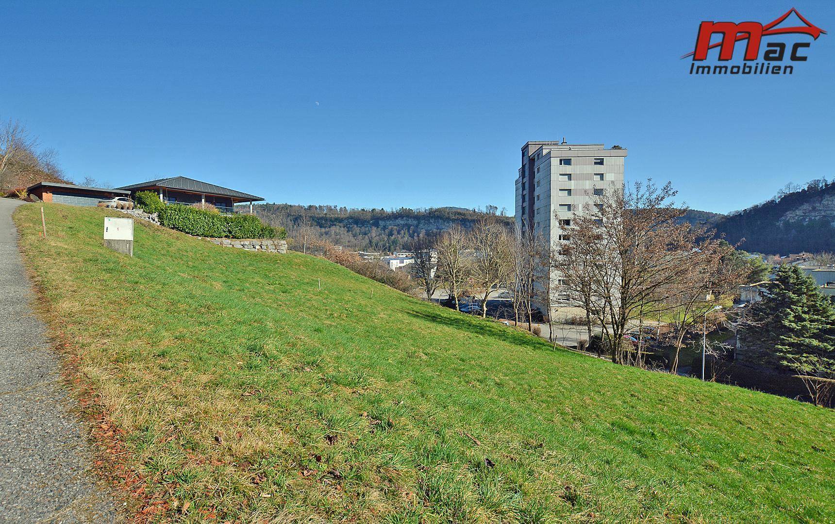 Grüne Hanglage mit einem Haus, einem hohen Gebäude und einem See in der Ferne unter blauem Himmel.