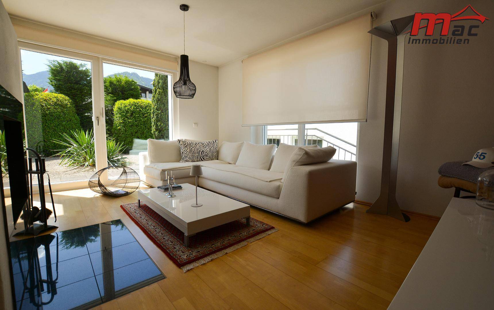 Gemütliches Wohnzimmer mit weißem Sofa, Holzboden und großen Fenstern mit Blick ins Grüne.