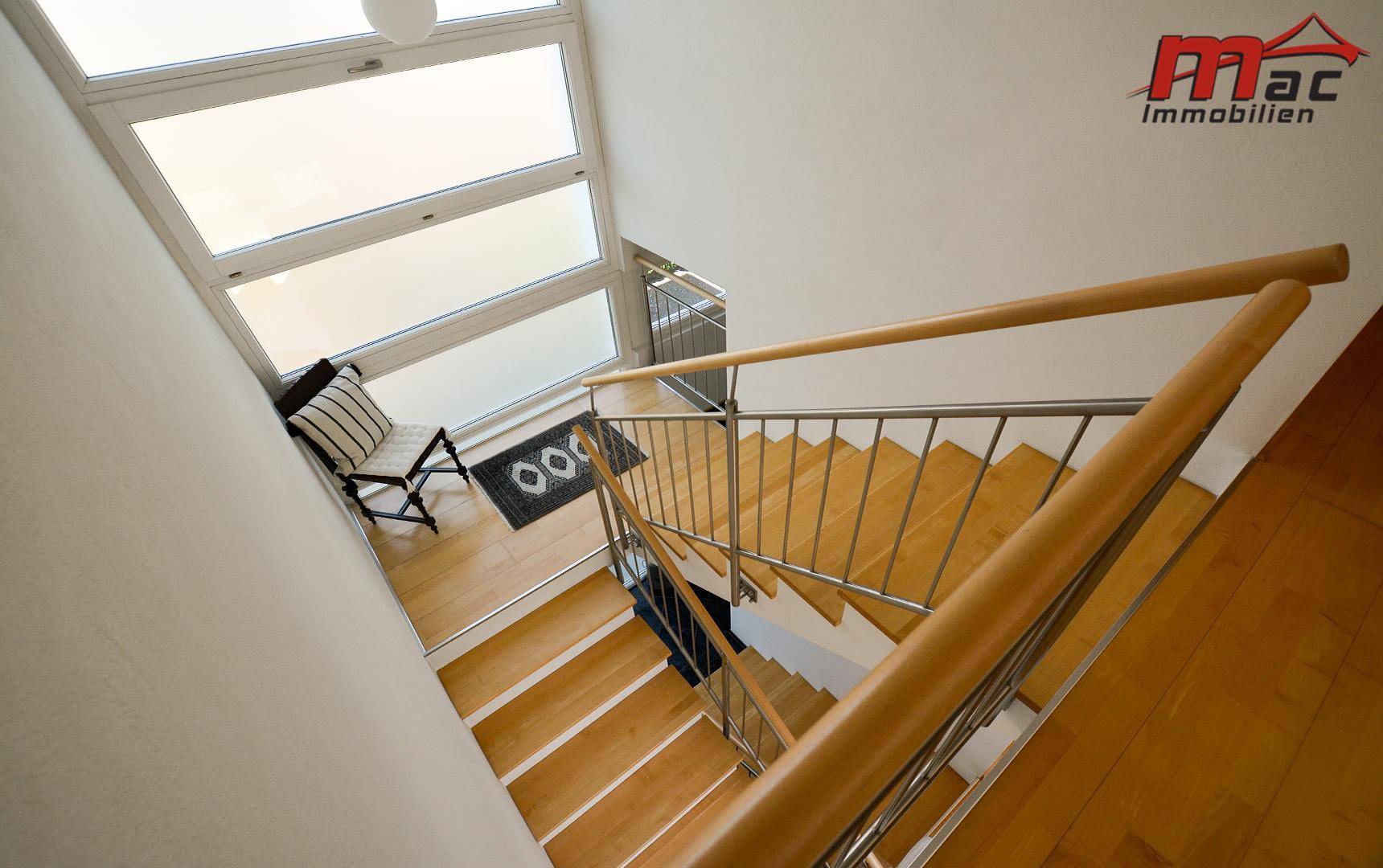 Moderne Holztreppe mit Edelstahlgeländer und großen Fenstern, die viel Licht hereinlassen.