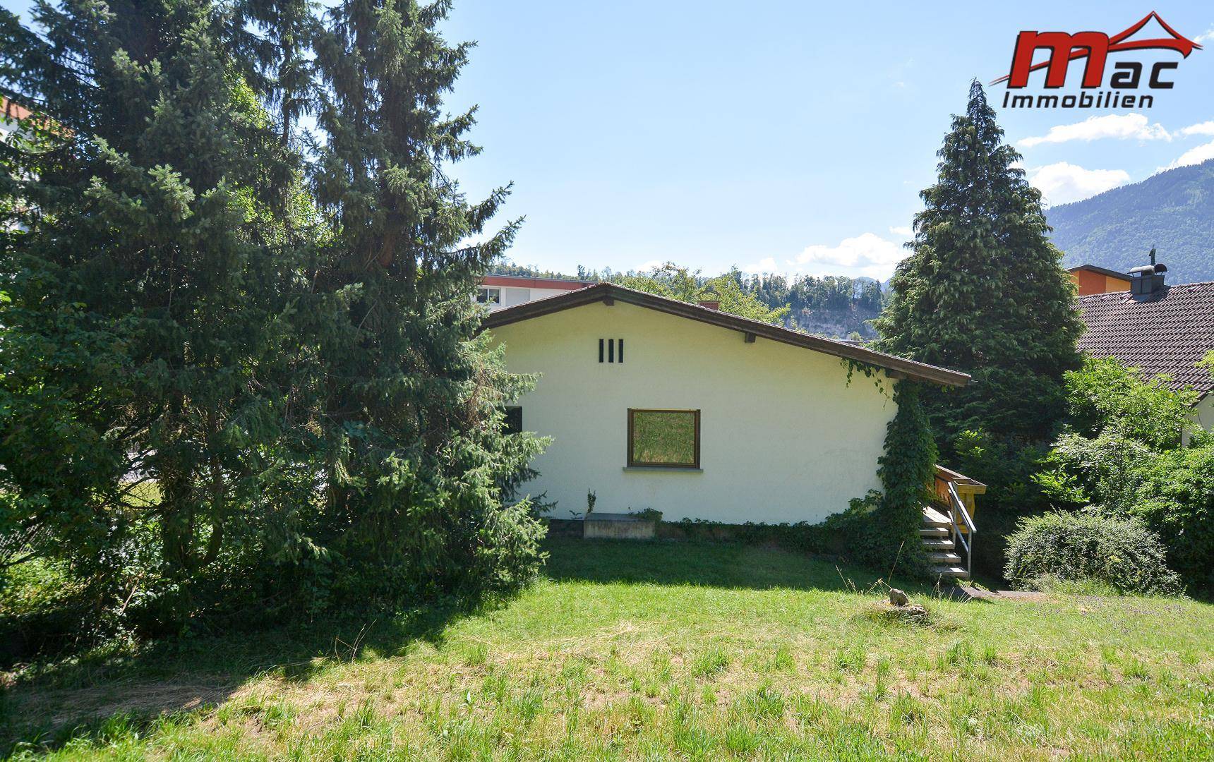 Rückseite des Hauses mit Rasenfläche, hohen Bäumen und Blick auf die umliegende Berglandschaft.