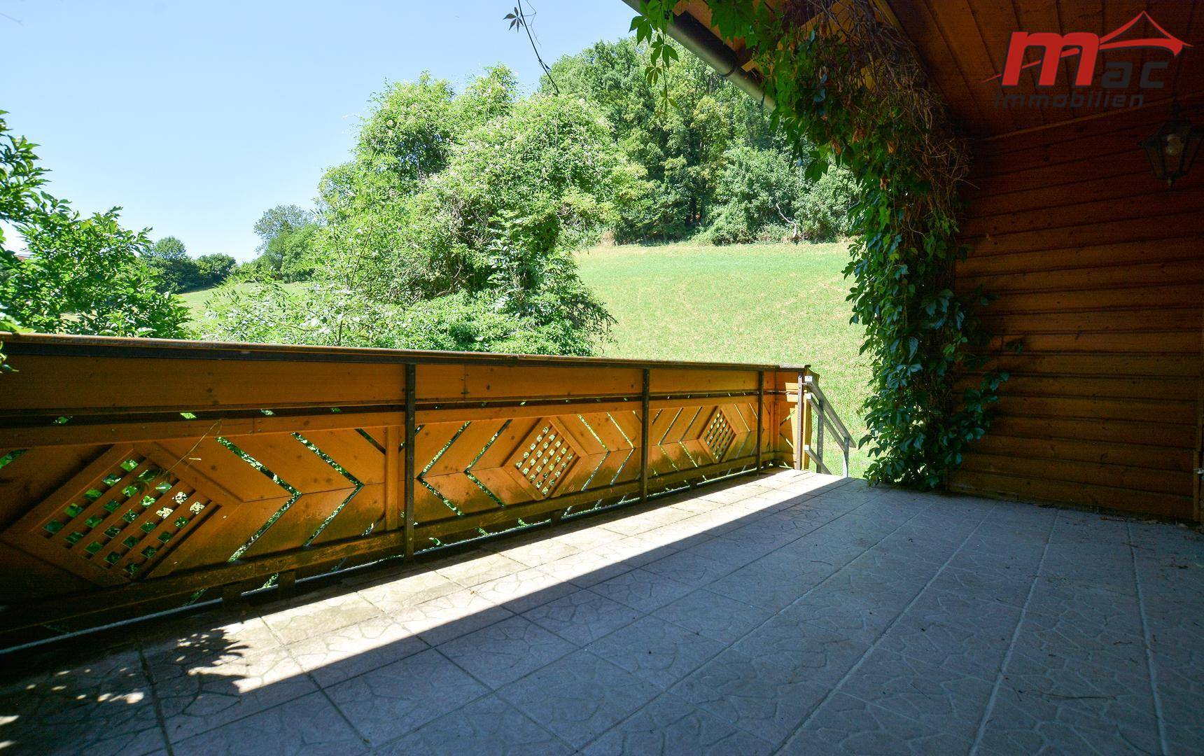 Sonniger Balkon mit Fliesenboden und Holzgeländer, bietet einen weiten Blick auf die grüne Landschaft.