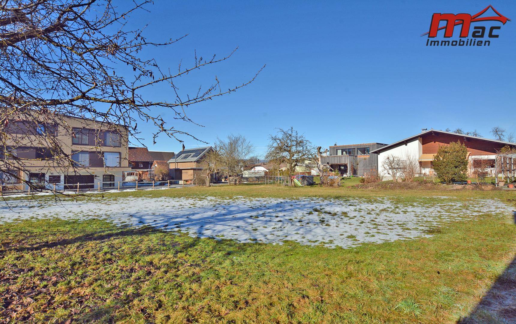 Offenes Grundstück mit grünen Flächen und Schneeresten, umgeben von Wohngebäuden unter blauem Himmel.