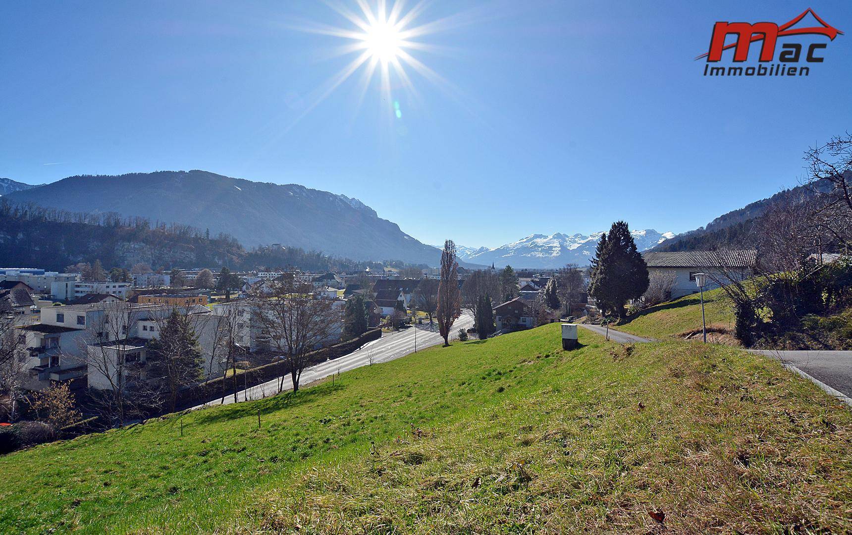Panoramablick über eine grüne Hügellandschaft mit einem Dorf und schneebedeckten Bergen im Hintergrund unter strahlendem Sonnenschein.