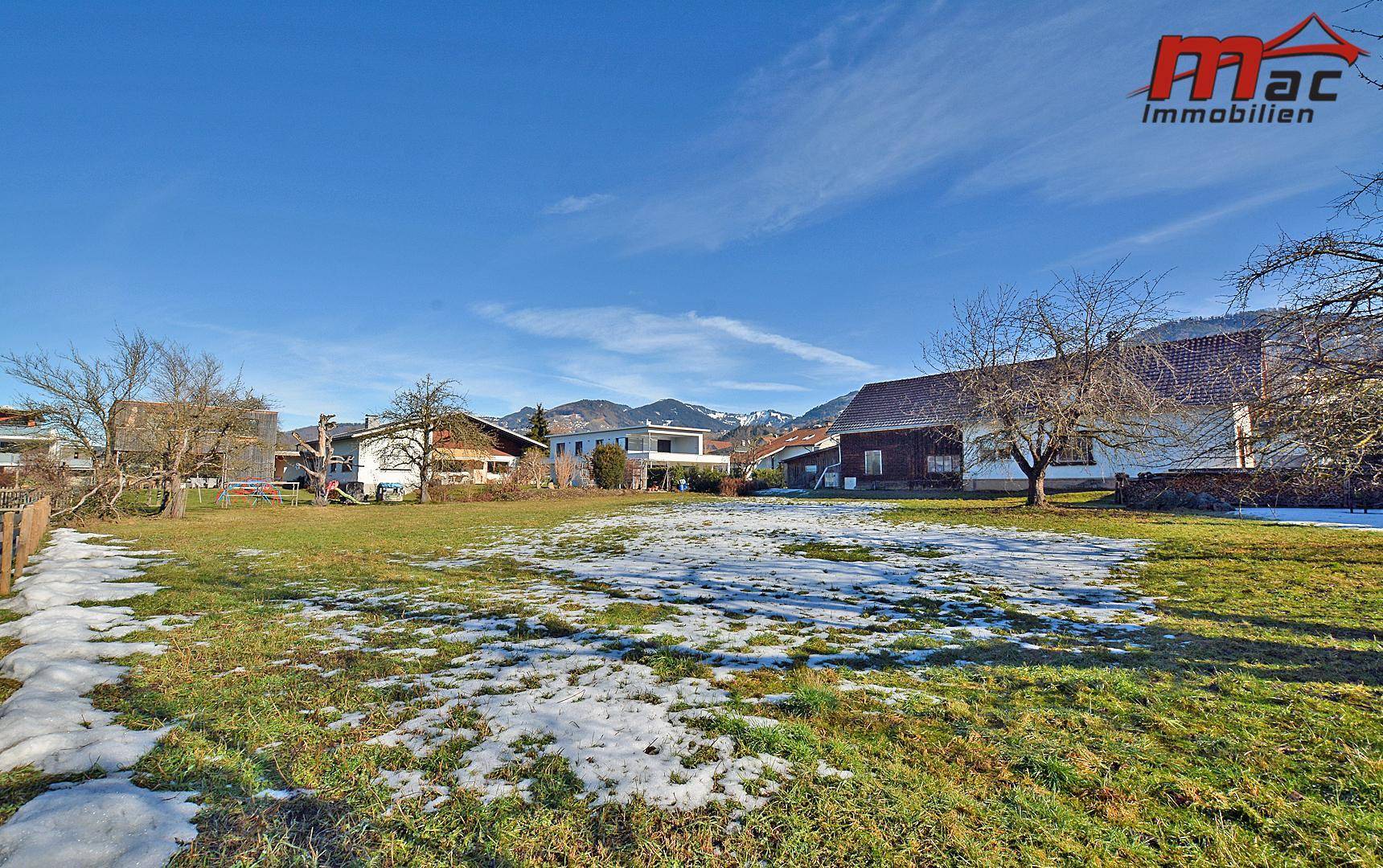 Weitläufiges Grundstück mit Gras und Schneeflecken, umgeben von verschiedenen Wohnhäusern und Bergpanorama.