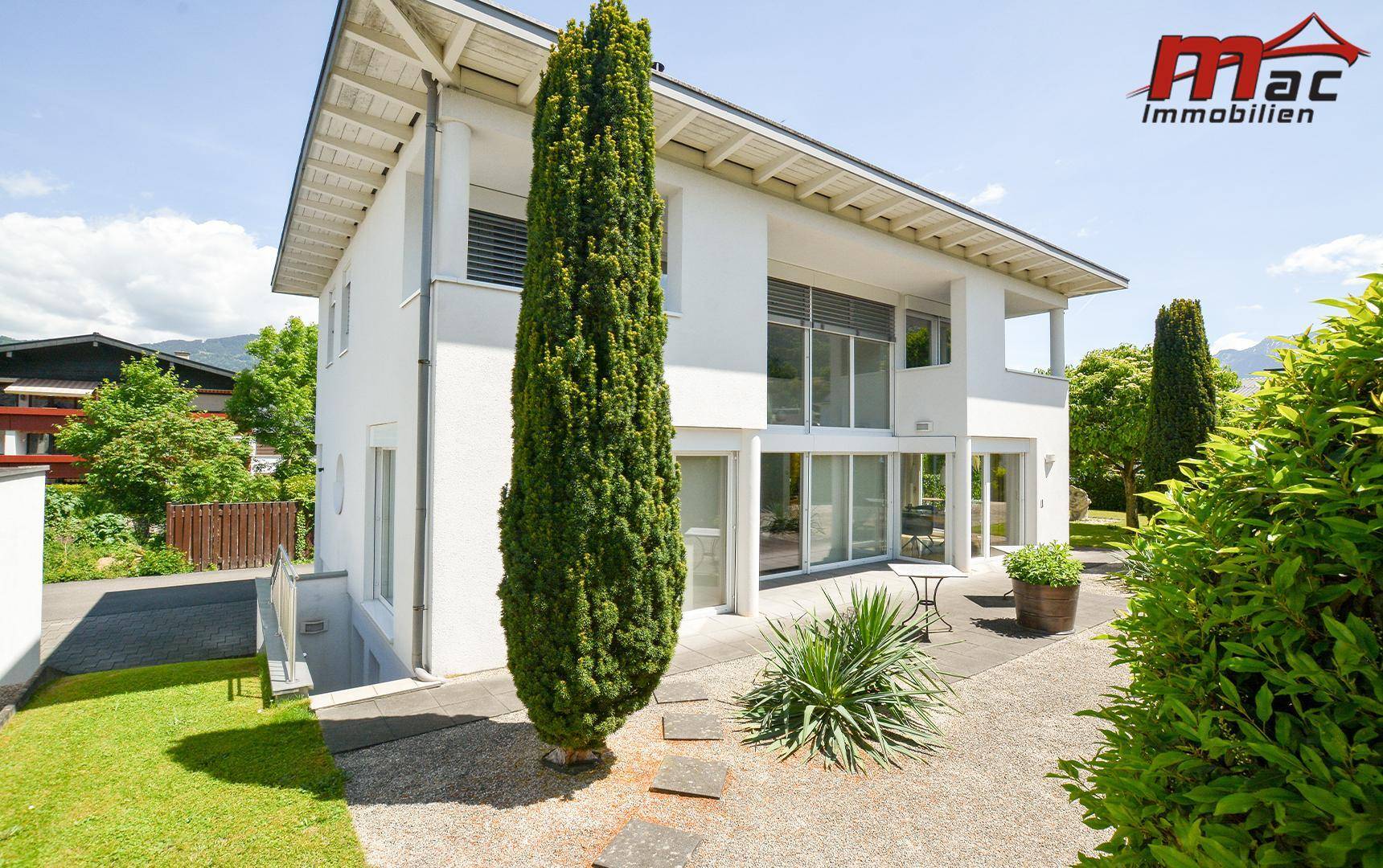 Weißes Einfamilienhaus mit großen Fensterfronten und gepflegtem Gartenbereich an einem sonnigen Tag.