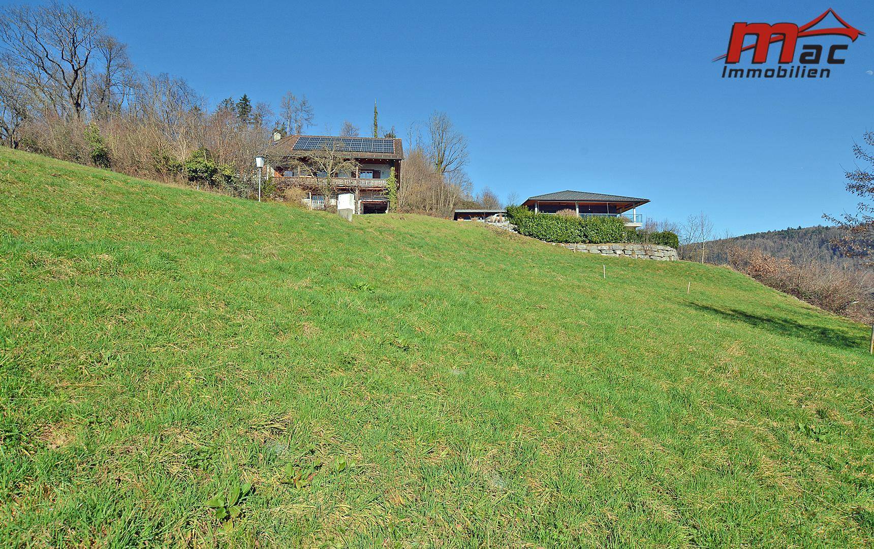 Weitläufige grüne Wiese auf einem Hang mit zwei Wohnhäusern in der Ferne, eines mit Solaranlage.