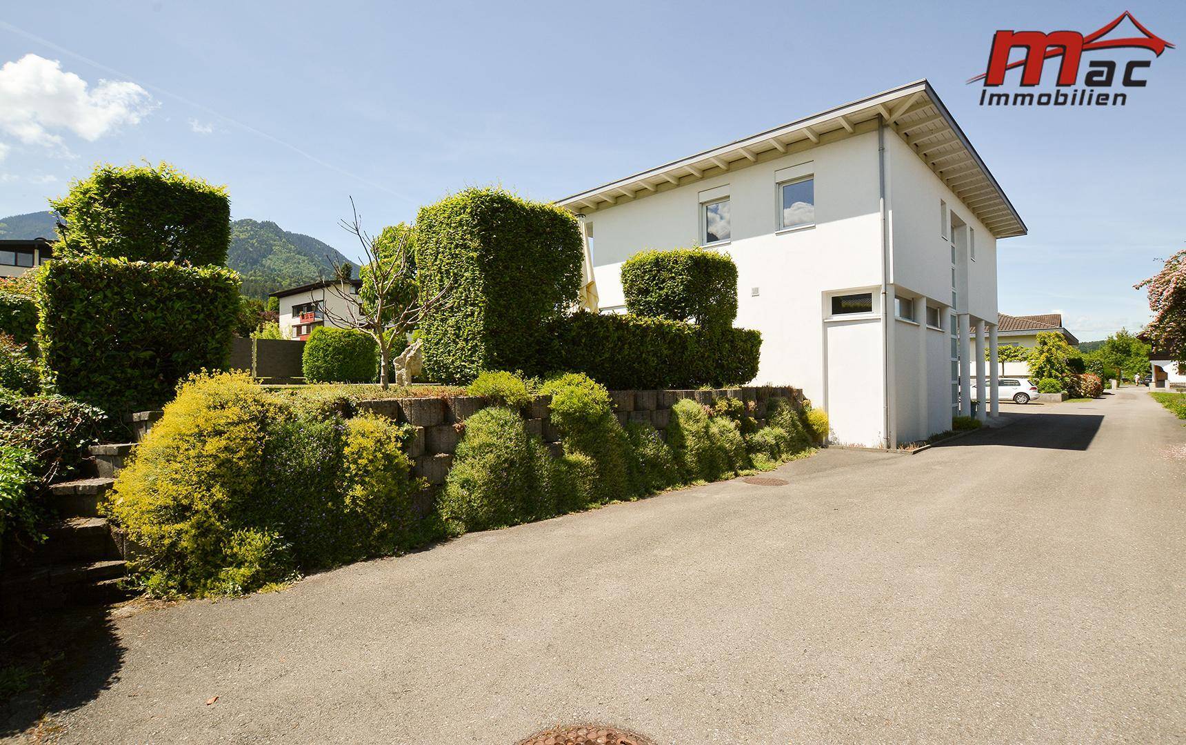 Modernes Einfamilienhaus mit gepflegtem Garten und Hecken, umgeben von grüner Landschaft.