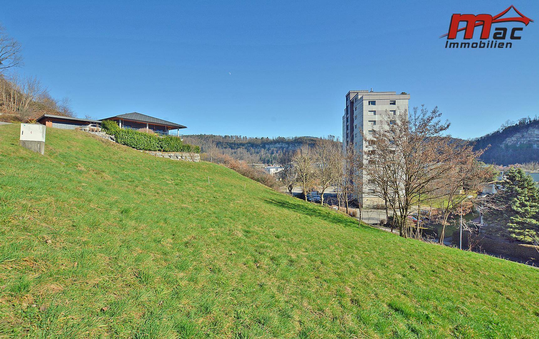 Grüne Hanglage mit einem Haus, einem hohen Gebäude und einem See in der Ferne unter blauem Himmel.