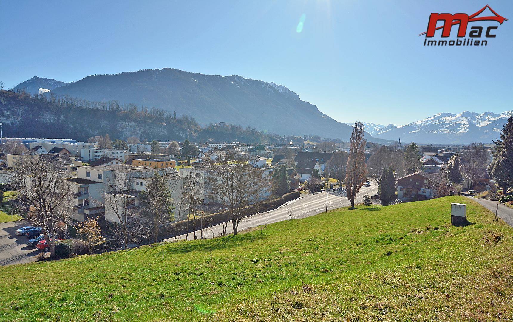 Panoramablick über eine grüne Hügellandschaft mit einem Dorf und schneebedeckten Bergen im Hintergrund unter strahlendem Sonnenschein.