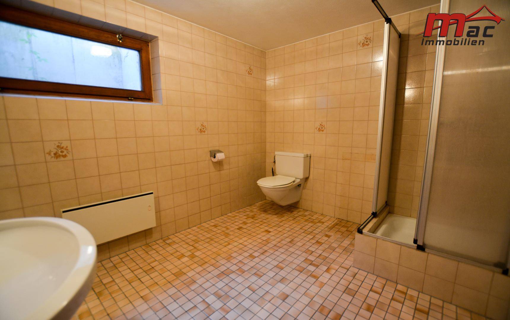 Gefliestes Badezimmer mit Dusche, Toilette, kleinem Waschbecken und einem Fenster für Tageslicht.