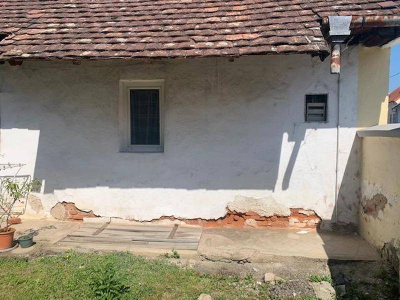Hauswand mit sichtbaren Putzschäden und einem kleinen Fenster, das Renovierungsbedarf anzeigt.