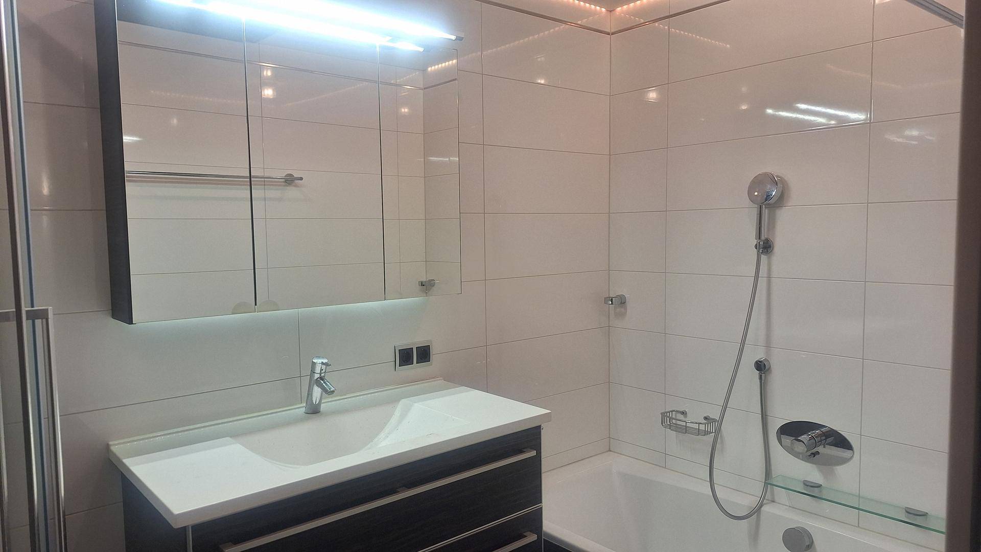 Modernes Badezimmer mit Waschtisch, Spiegelschrank und Badewanne mit Duschfunktion, hell gefliest.