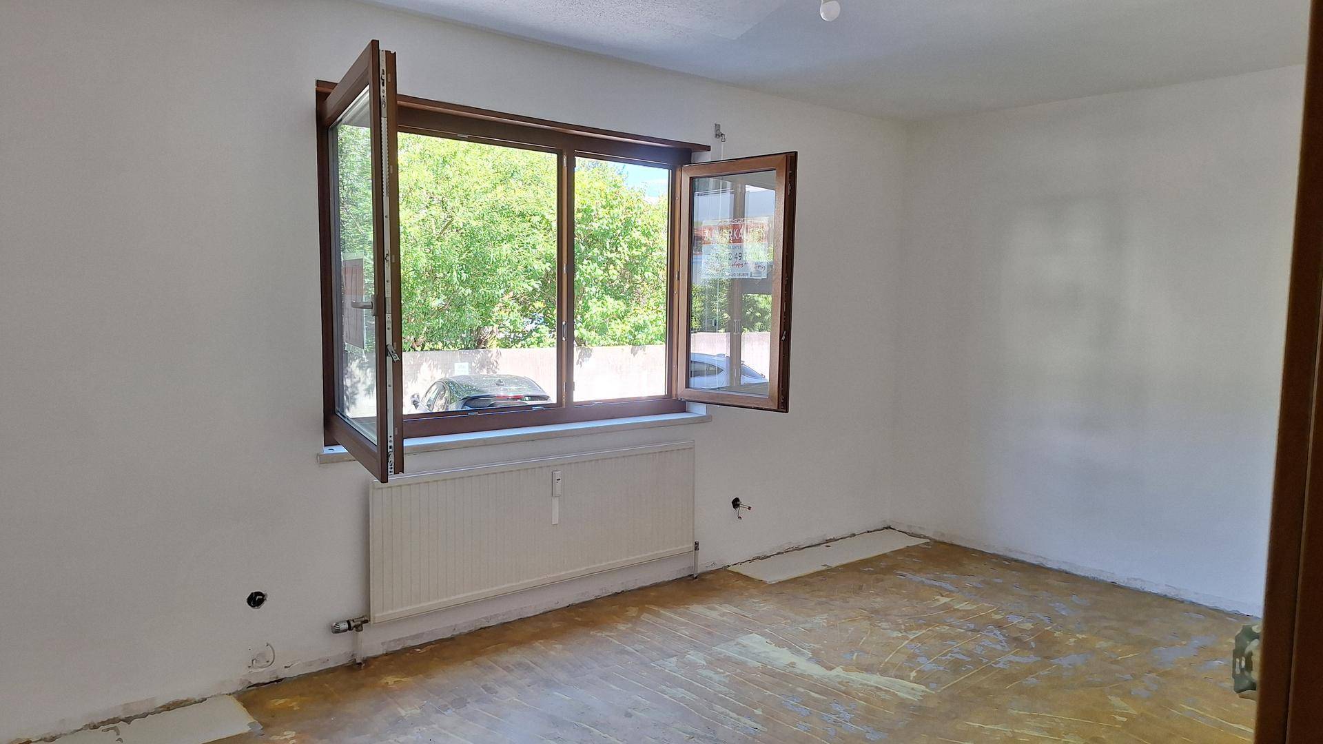 Leeres Zimmer mit großen Fenstern, die einen Blick auf Bäume und die Umgebung bieten.